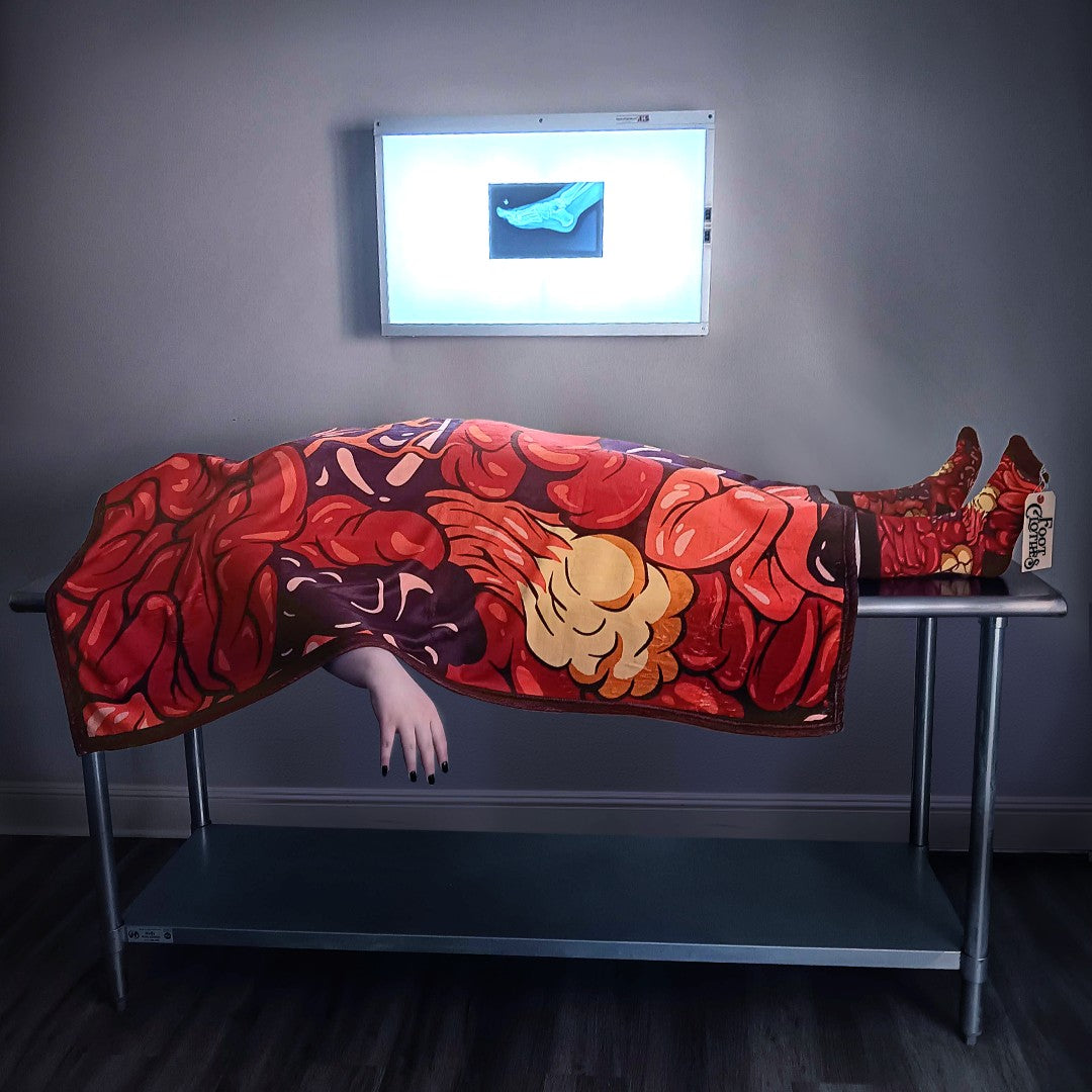A person lays on a medical table covered from head to toe in FootClothes Gut Feeling Blanket and matching socks with toe tag in front of an Xray viewer. The image indicates the person is deceased in a humorous manner.
